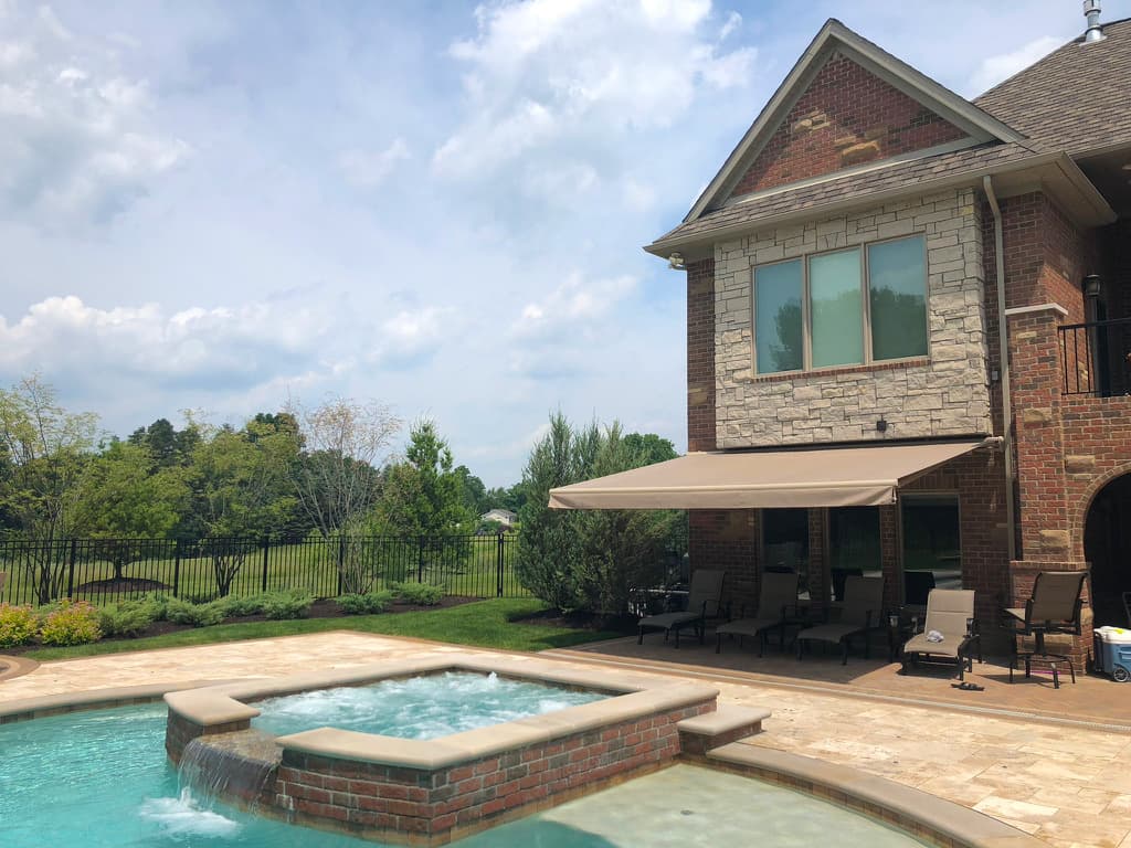 Beige retractable awning shading a brick home's patio beside an in-ground pool